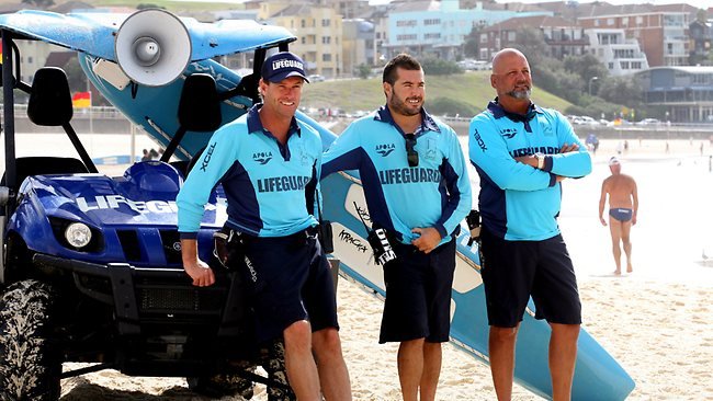 Three Bondi Rescue lifeguards in teal uniforms standing beside a rescue vehicle with surfboards on Bondi Beach